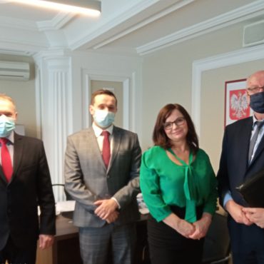 Grupa trzech mężczyzn w garniturach wraz z kobietą znajdują się w pomieszczeniu biurowym.Obok nich stoi biurko, na ścianie w kolorze bezowym wisi godło Polski.