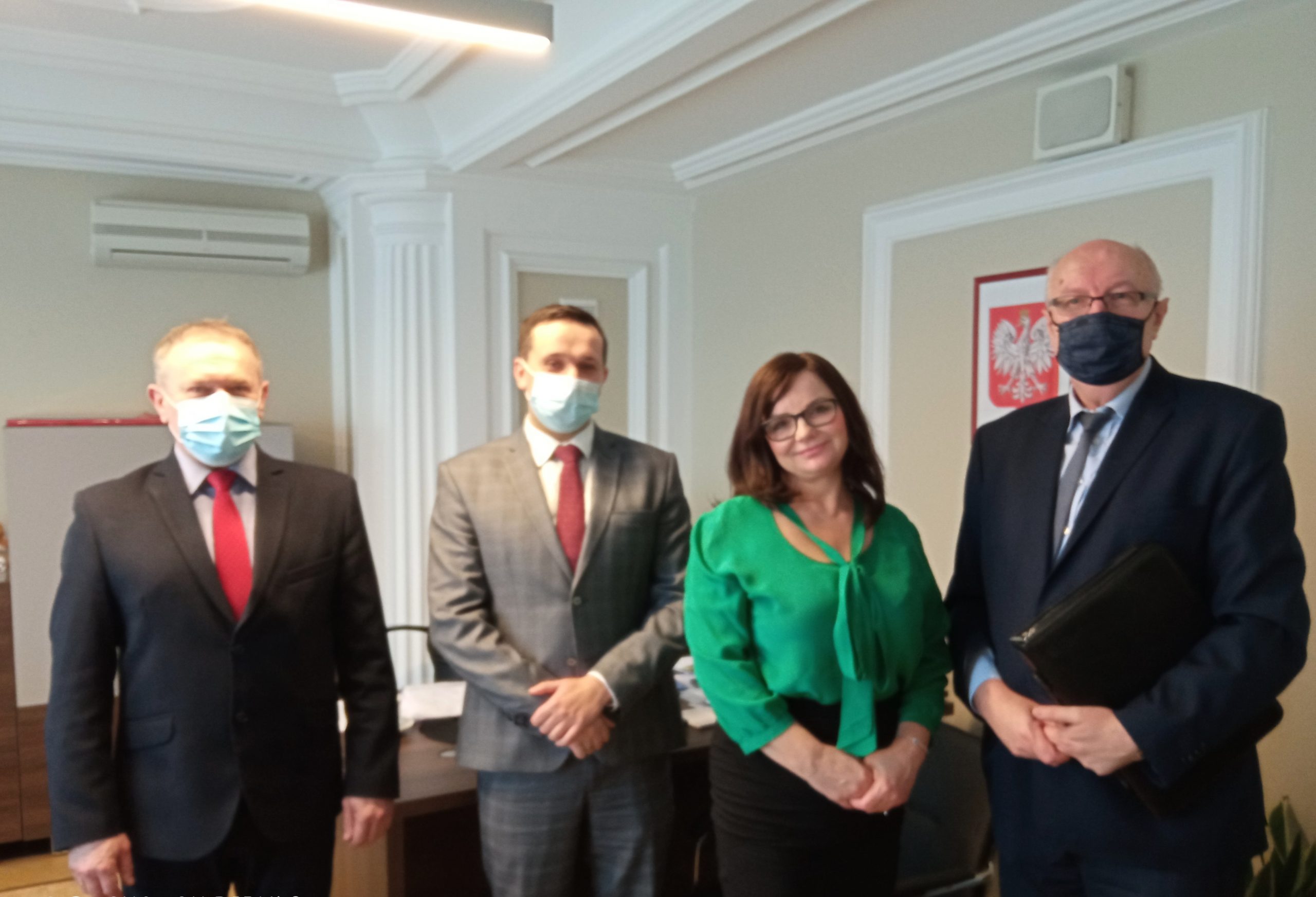 Grupa trzech mężczyzn w garniturach wraz z kobietą znajdują się w pomieszczeniu biurowym.Obok nich stoi biurko, na ścianie w kolorze bezowym wisi godło Polski.