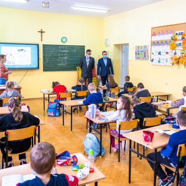 Dwóch mężczyzn w garniturach przebywa w pomieszczeniu z dziećmi, które siedzą przy stolikach.