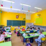 Grupa kilkudziesięciu dzieci siedzących przy stolikach podczas lekcji.