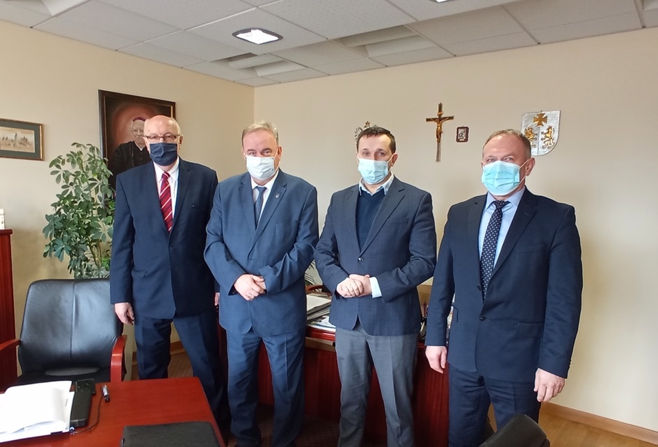 Czterech mężczyzn w garniturach i maseczkach stoją w jasnym pomieszczeniu.