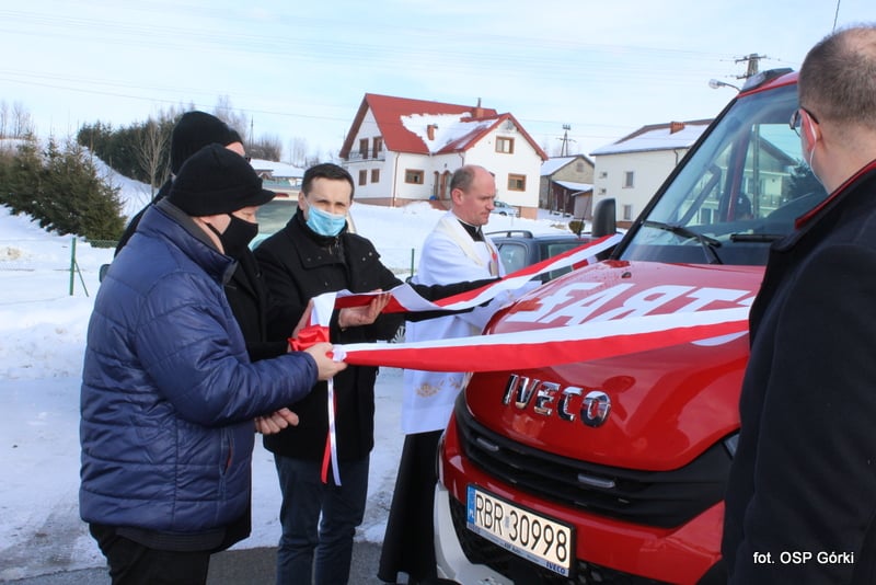 Trzech mężczyzn, w tym ksiądz przecina czerwono - białą wstęgę na samochodzie pożarniczym.