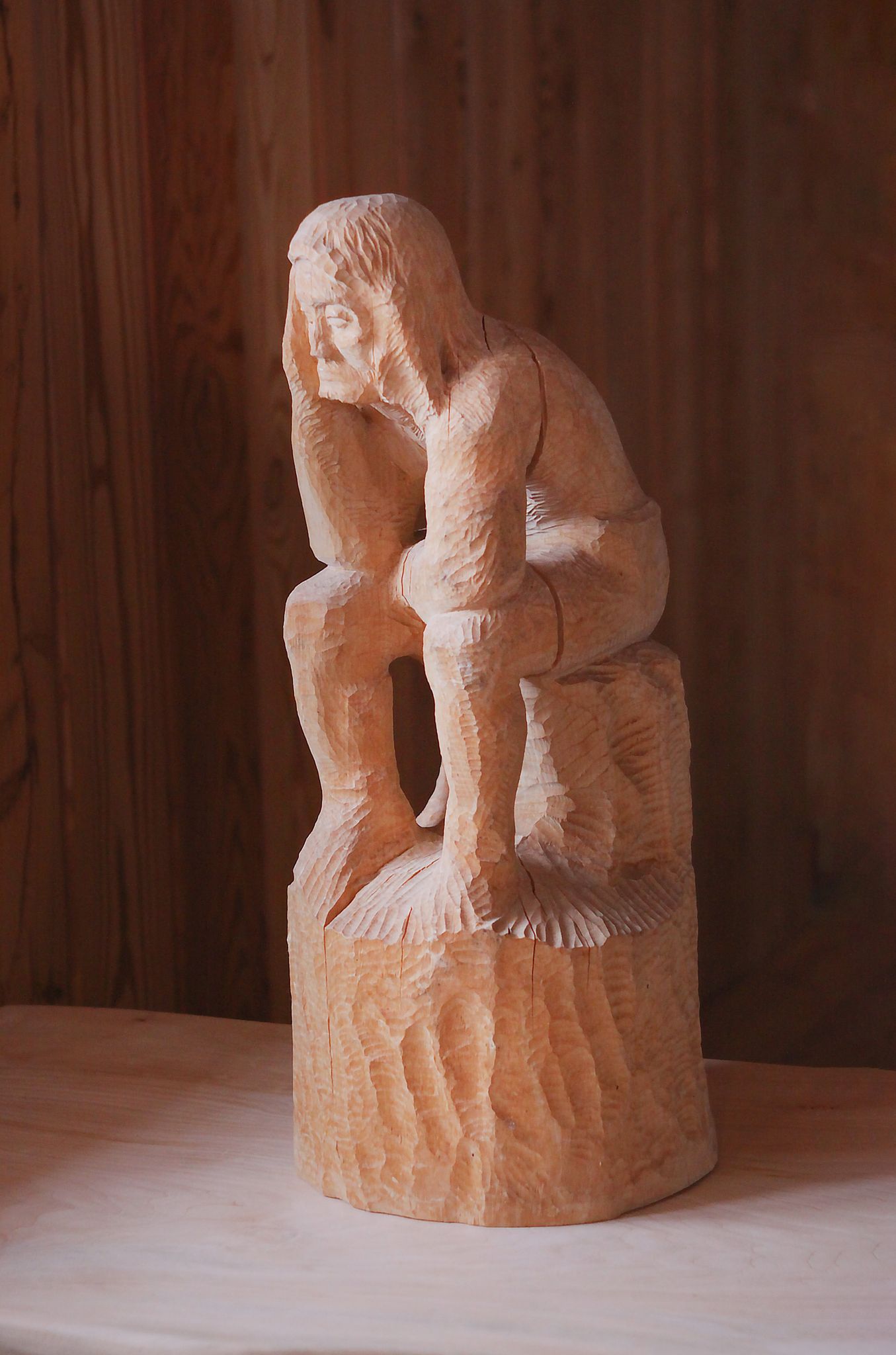 Figurka z drewna przedstawiająca mężczyznę siedzącego na pniu z trzymającymi ręce na głowie.