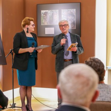 Starszy mężczyzna w garniturze przemawia do publiczności zgromadzonej w muzeum. Obok mężczyzna stoi kobieta w zielonej sukience i czarnej marynarce. Pomieszczenie w jasnych kolorach, w którym wystawione są fotografie.