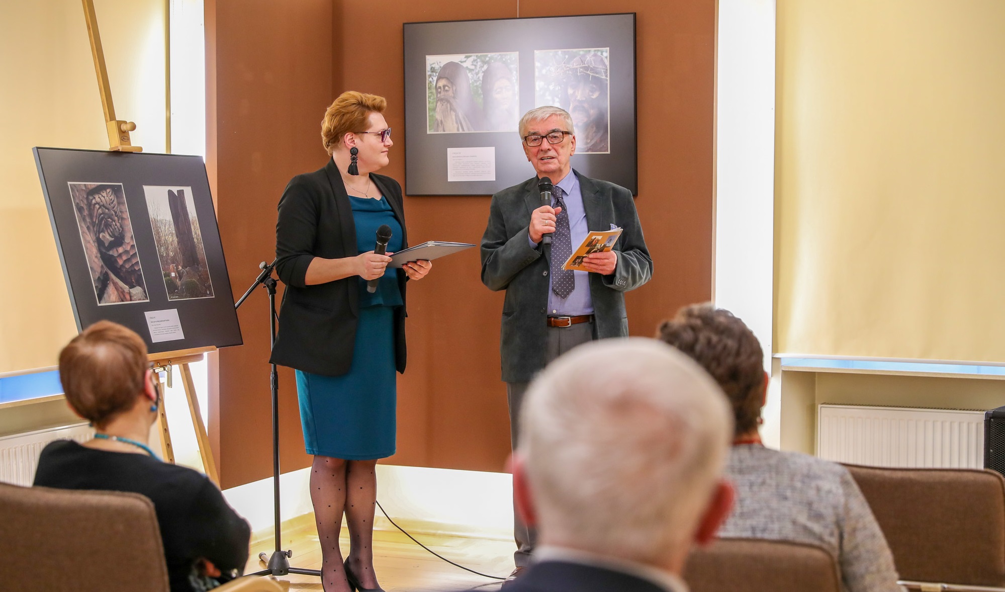 Starszy mężczyzna w garniturze przemawia do publiczności zgromadzonej w muzeum. Obok mężczyzna stoi kobieta w zielonej sukience i czarnej marynarce. Pomieszczenie w jasnych kolorach, w którym wystawione są fotografie.