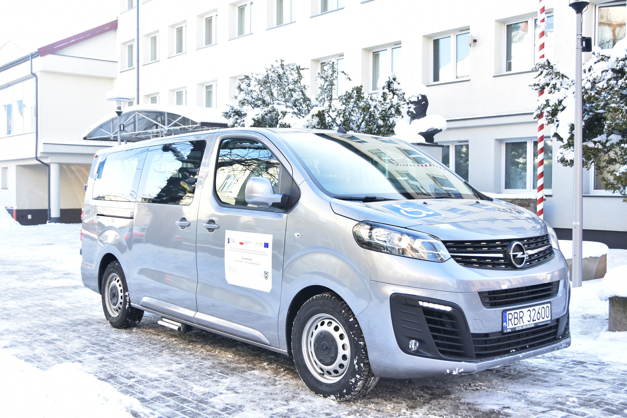 Samochód marki Opel koloru szarego stoi zaparkowany przed białym piętrowym budynkiem.