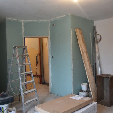 Pomieszczenie wewnątrz budynku. Trwają w nim prace remontowe. Są brudne ściany, podłoga nie ma paneli, postawione są ścianki, z których powstanie łazienka.