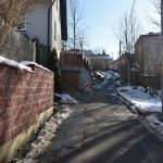 Droga asfaltowa prowadzi do budynków mieszkalnych. Otoczona z jednej strony skarpą wyłożoną cegła, a z drugiej strony budynkiem.