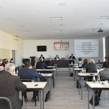 Grupa osób siedzi przy stolikach w jasnym dużym pomieszczeniu. Przed nimi na ścianie wisi telewizor oraz wyświetlane są informacje projektorem.