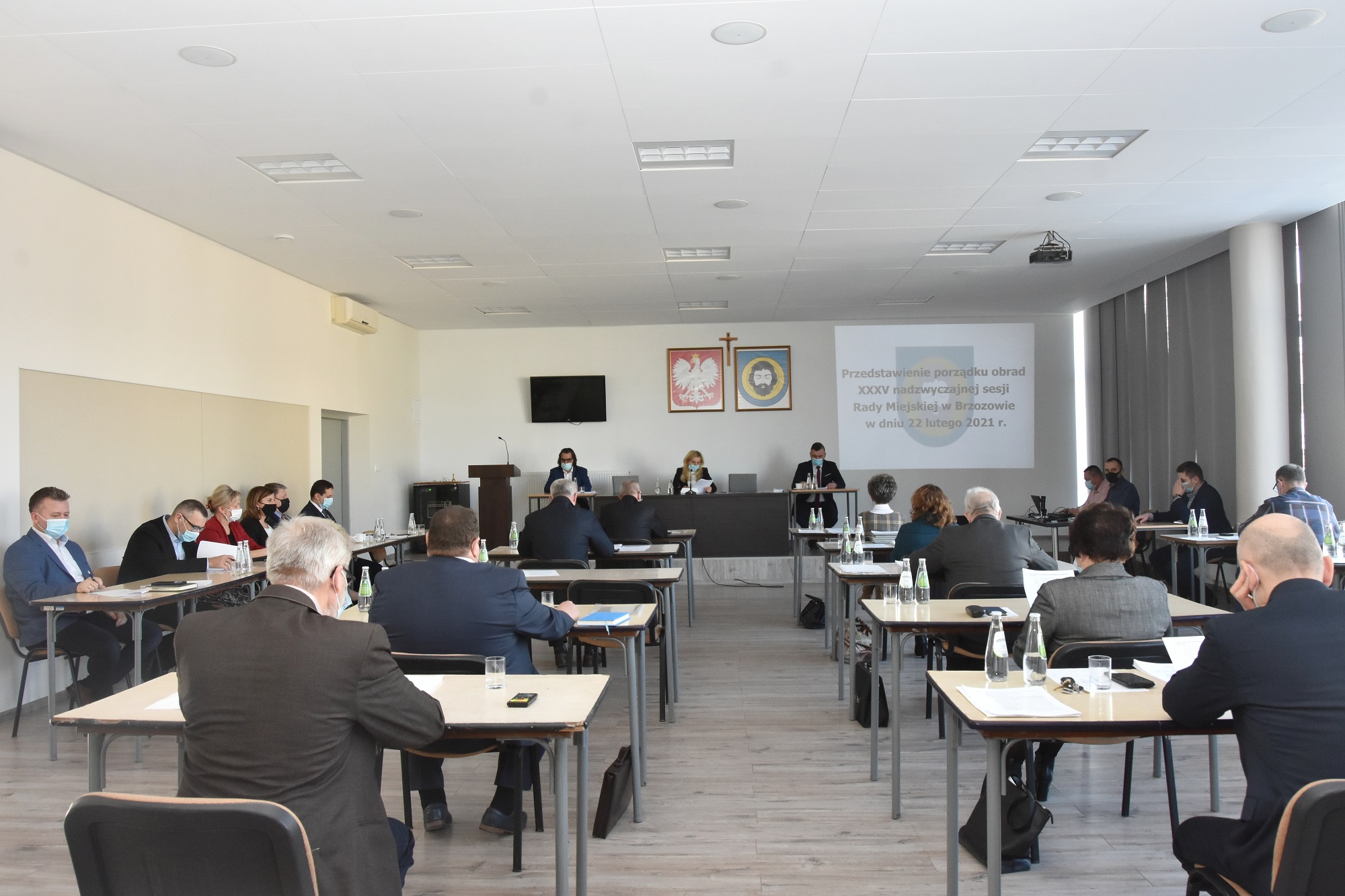 Grupa osób siedzi przy stolikach w jasnym dużym pomieszczeniu. Przed nimi na ścianie wisi telewizor oraz wyświetlane są informacje projektorem.