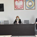 Grupa trzech osób siedzi przy stoliki. Za nimi na ścianie wisi telewizor i obrazy przedstawiające godło Polski i herb Gminy.