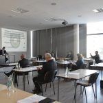 Grupa osób z podniesioną ręką siedzi przy stolikach w jasnym pomieszczeniu z dużymi oknami.