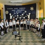 Grupa dzieci ubranych odświętnie stoi w półkolu i śpiewa piosenki. Na środku stoi mikrofon.