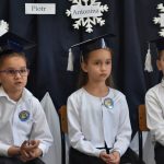 Dwie dziewczynki i chłopczyk w białych koszulach i papierowych czapkach na głowie siedzą na krzesełkach.