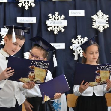 Dzieci ubrane w białe koszule stoją na korytarzu i trzymają w rękach granatowe dyplomy.