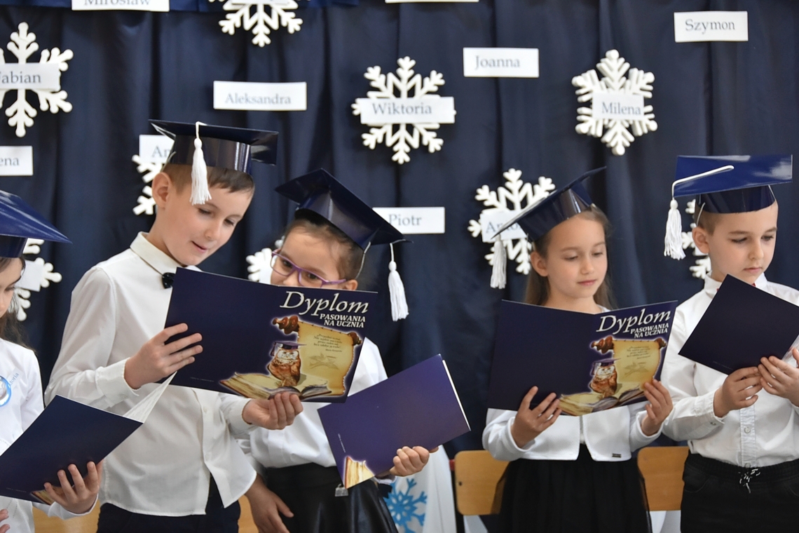 Dzieci ubrane w białe koszule stoją na korytarzu i trzymają w rękach granatowe dyplomy.
