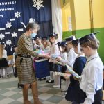 Kobieta w sukience w kratkę przekazuje dzieciom malowanki. Dzieci stoją na korytarzu, ściany koloru zielono-żóltego, podłoga wyłożona płytkami.