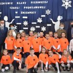 Grupa dzieci w pomarańczowych koszulkach, z lewej strony stoi dwóch mężczyzn w garniturach i kobieta w brązowej marynarce, z prawej strony stoją dwie kobiety jedna w kratkę w sukience, druga w pomarańczowej koszulce, a między nimi mężczyzna w garniturze. Wszyscy stoją przed ciemną tablicą na której przywieszone są białe literki.