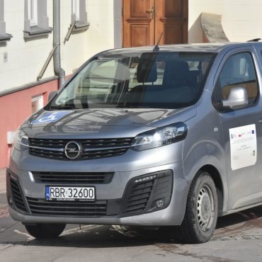 Samochód koloru szarego zaparkowany przed duzym budynkiem jasnej elewacji w zimowej odsłonie.