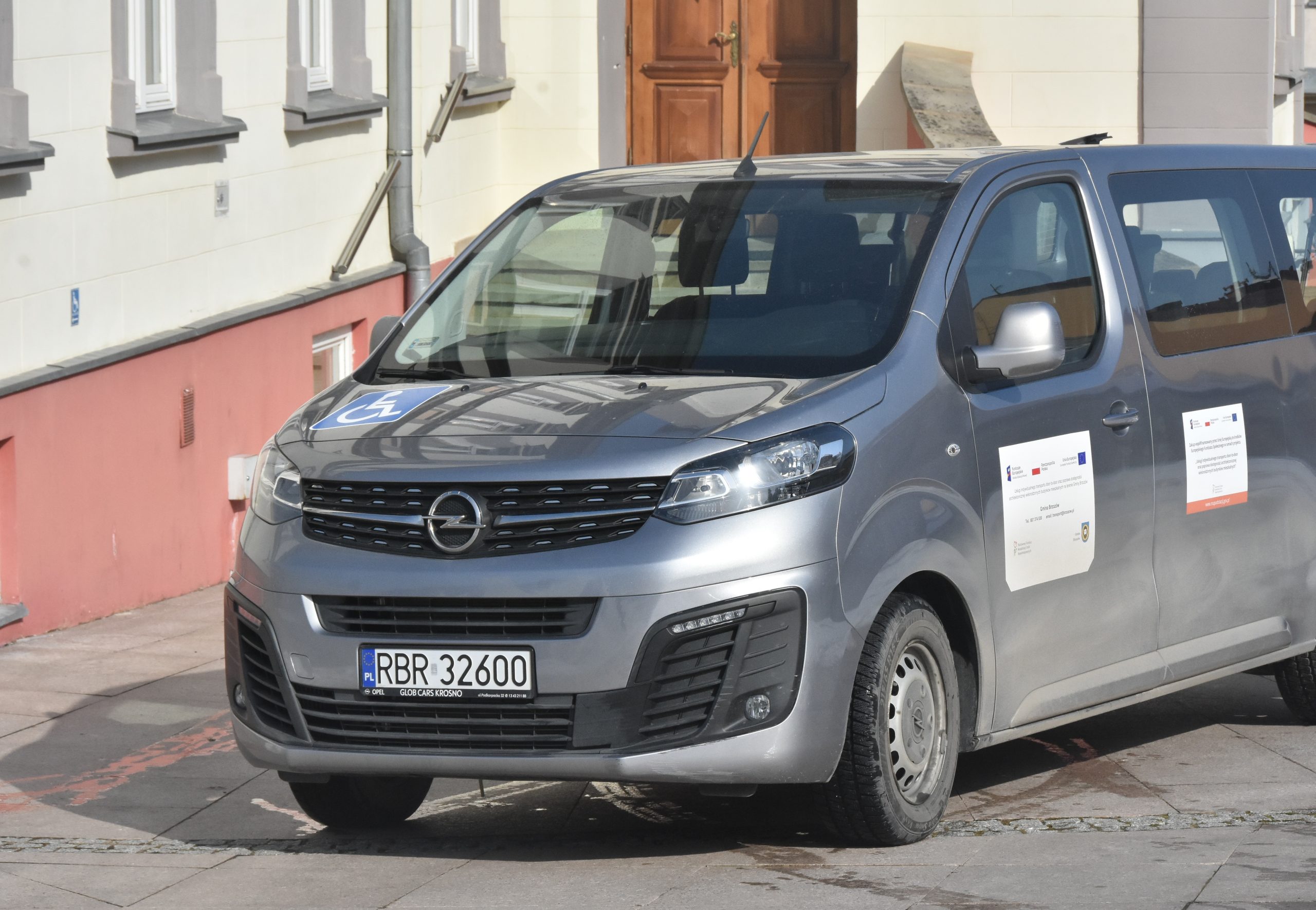 Samochód koloru szarego zaparkowany przed duzym budynkiem jasnej elewacji w zimowej odsłonie.