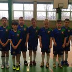 Grupa kilkudziesięciu chłopców w różnokolorowych strojach piłkarskich, stoją na sali gimnastycznej. Obok nich stoi dwóch trenerów.