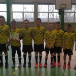 Grupa kilkudziesięciu chłopców w różnokolorowych strojach piłkarskich, stoją na sali gimnastycznej. Obok nich stoi trzech trenerów.