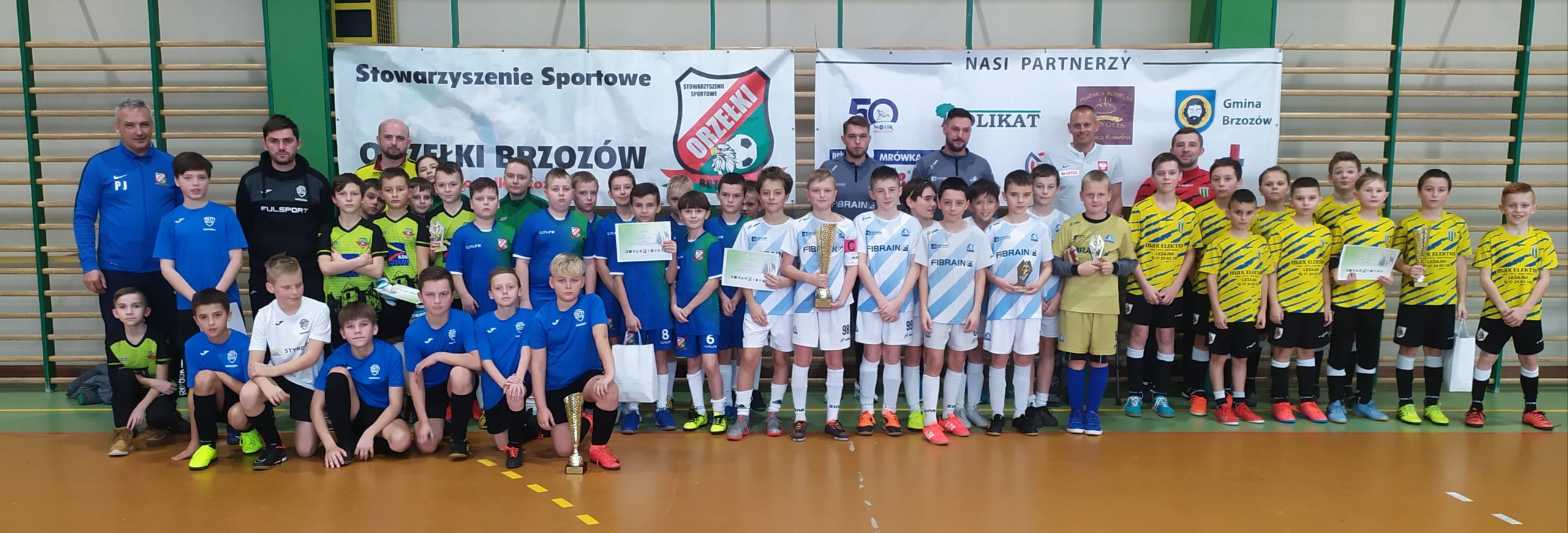 Grupa kilkudziesięciu dzieci ubranych w stroje piłkarskich stoją na sali gimnastycznej. W centrum stoi chłopak trzymający puchar.