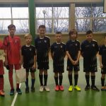 Grupa kilkudziesięciu chłopców w różnokolorowych strojach piłkarskich, stoją na sali gimnastycznej. Obok nich stoi trzech trenerów.