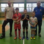 Grupa trzech dzieci ubranych w stroje piłkarskich stoją na sali gimnastycznej. Chłopcy trzymają statuetki, obok nich stoją trenerzy.