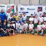 Grupa kilkudziesięciu dzieci ubranych w stroje piłkarskich stoją na sali gimnastycznej. W centrum stoi chłopak trzymający puchar.