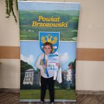 Dziewczynka w czarnych leginsach i białej bluzce trzyma w rękach dyplom i zeszyt reklamowy. Dziewczynka stoi przed banerem Powiatu Brzozowskiego.