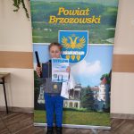 Chłopczyk w jasnych włosach i ciemnym swetrze stoi przed banerem powiatu brzozowskiego i trzyma w rękach dyplom i gadżety promocyjne, tj. opaskę odblaskową i jasny zeszyt.