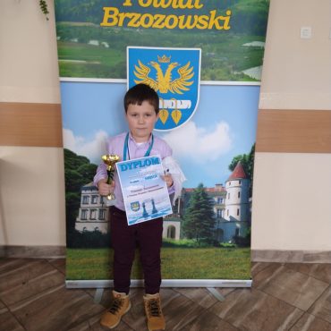 Mały chłopczyk trzymający w jednej ręce puchar, a w drugiej dyplom. Stoi przed banerem Powiatu Brzozowskiego.