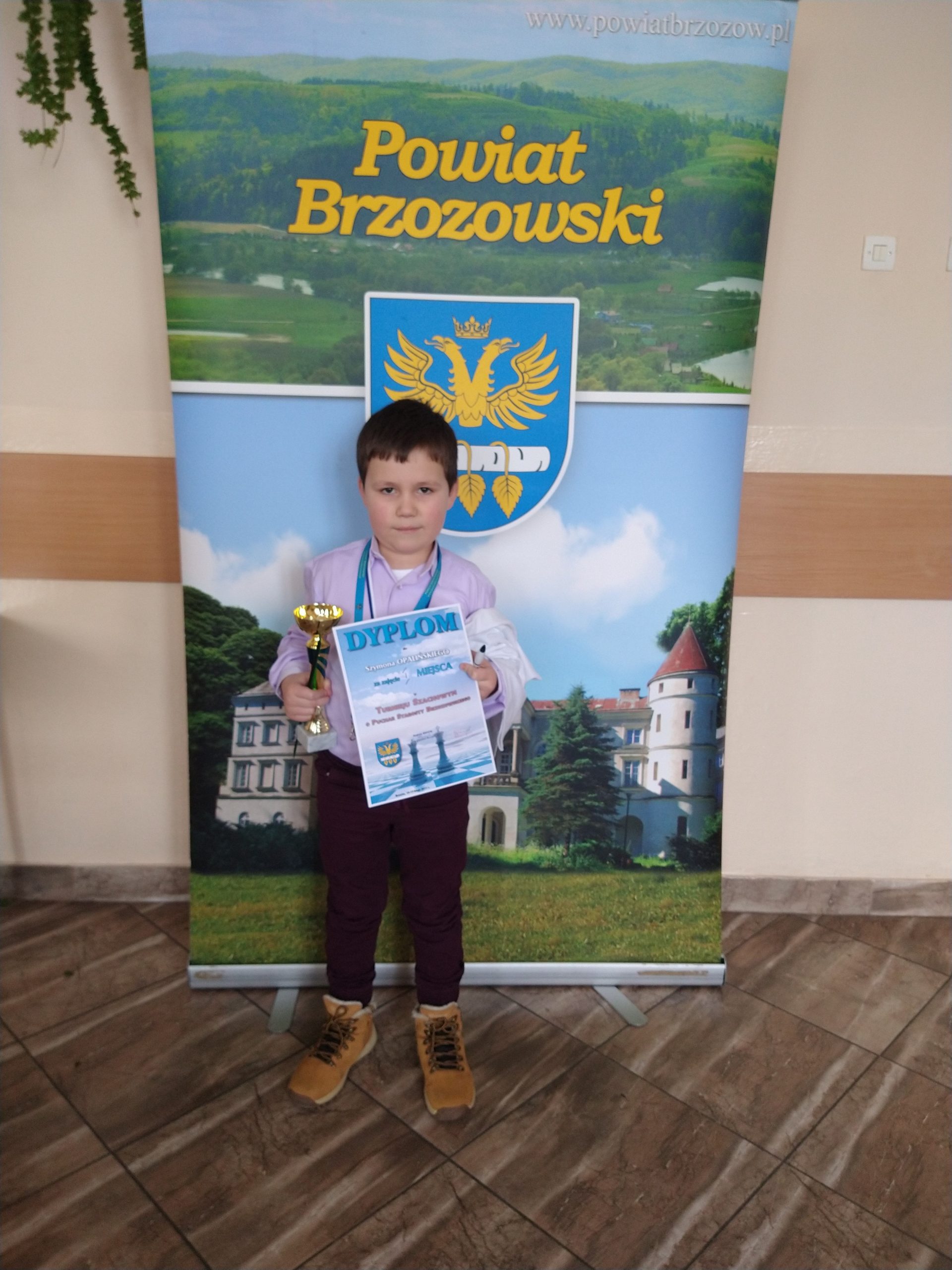 Mały chłopczyk trzymający w jednej ręce puchar, a w drugiej dyplom. Stoi przed banerem Powiatu Brzozowskiego.