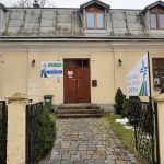 Stary budynek z kremową elewacją. Na środku drzwi w kolorze brązowym. Budynek otoczony bramą.