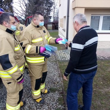 Dwóch mężczyzn w jasnych mundurach przekazują ulotkę starszemu mężczyźnie.