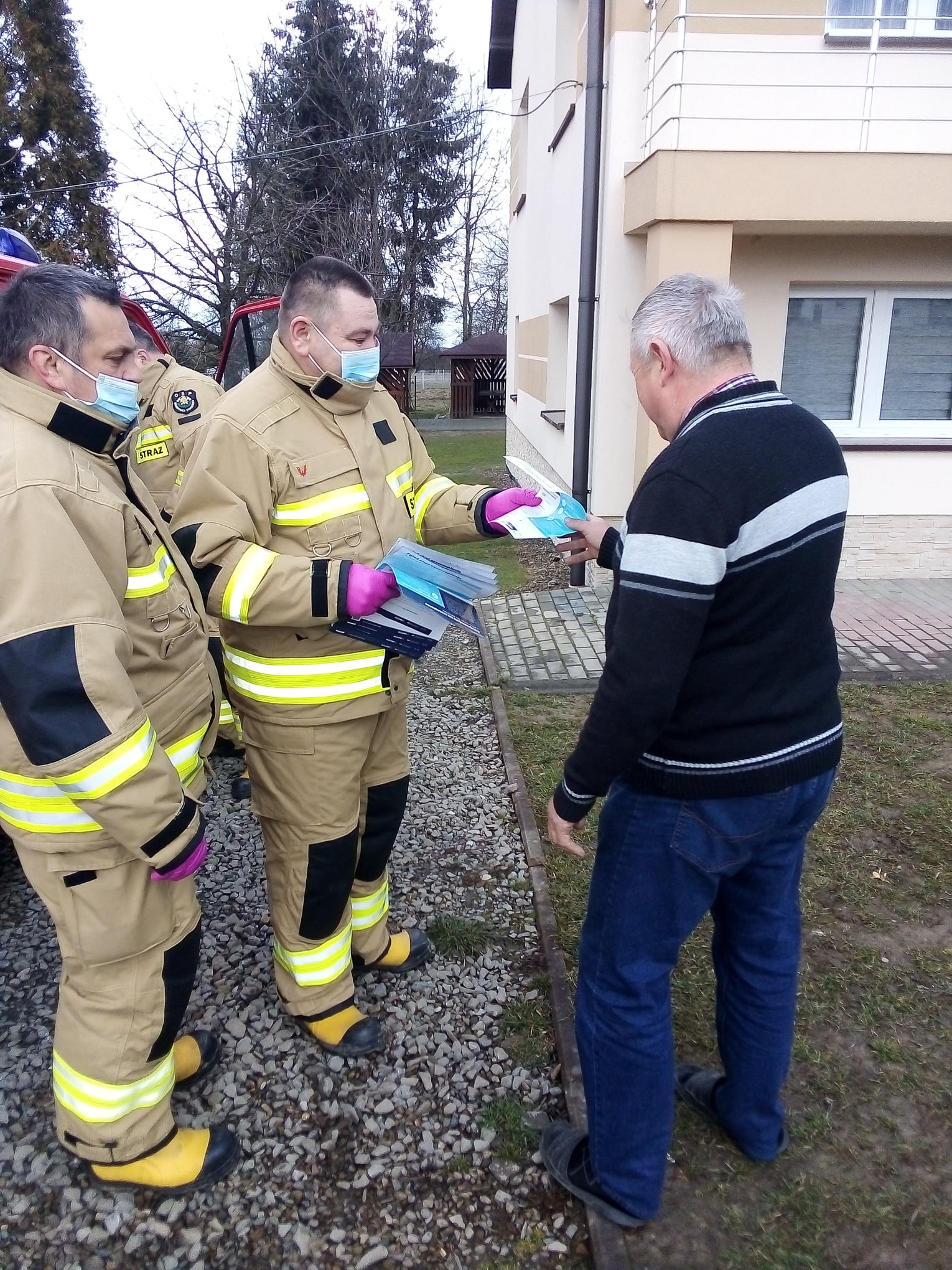 Dwóch mężczyzn w jasnych mundurach przekazują ulotkę starszemu mężczyźnie.