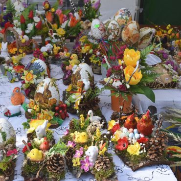 Na stole wyłożonym białym obrusem leżą różnorodne kolorowe pisanki, kurczaki wielkanocne, koszyki, stroiki, książeczki , palmy.