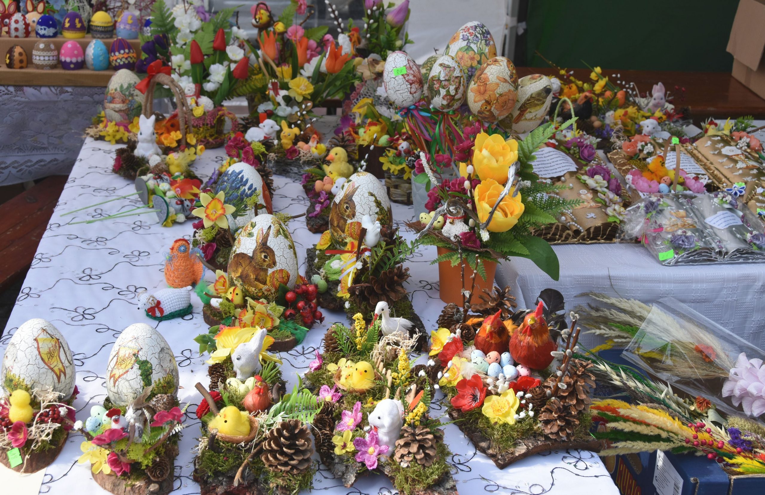 Na stole wyłożonym białym obrusem leżą różnorodne kolorowe pisanki, kurczaki wielkanocne, koszyki, stroiki, książeczki , palmy.