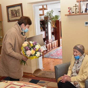 Starsza kobieta siedzi na fotelu, młoda kobieta trzyma kwiaty i czyta życzenia.