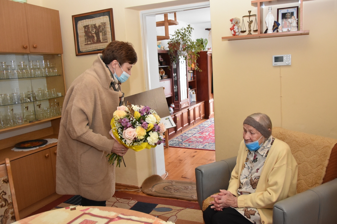 Starsza kobieta siedzi na fotelu, młoda kobieta trzyma kwiaty i czyta życzenia.