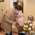 Młoda kobieta w jasnym płaszczu składa życzenia wraz z kwiatami starszej kobiecie, która siedzi w fotelu.