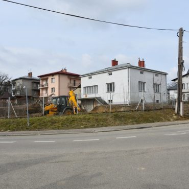 Plac w budowie. Na środku stoi koparka, przed bramą pomarańczowa ciężarówka.