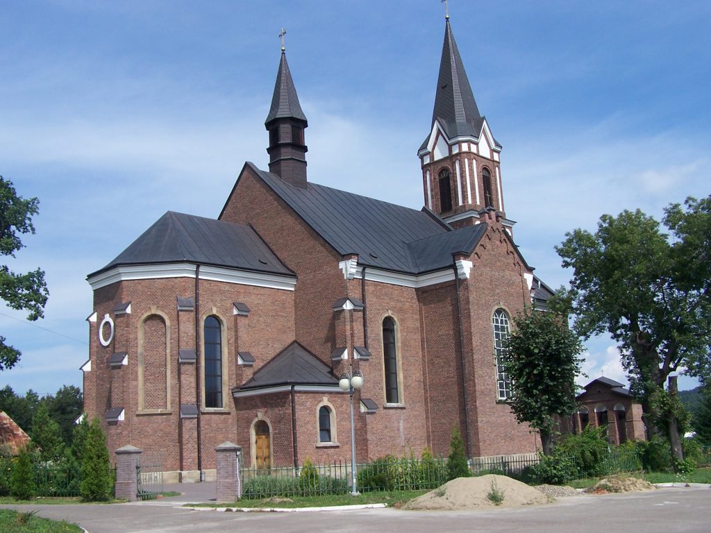 Kościół z ciemnej cegły, dach z szarociemnej blachy, kościół ogrodzony, przed kościołem drzewa liściaste.
