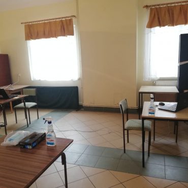 Jasne pomieszczenie w budynku. Stoją w nim stoliki, na których leżą laptopy.