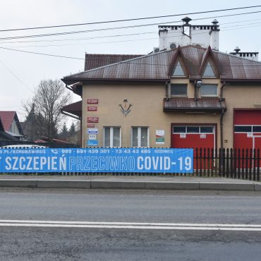 Budynek straży pożarnej z jasną elewacją, z dużymi drzwiami koloru czerwonego. Na ogrodzeniu przed budynkiem rozwieszony duży plakat koloru niebieskiego dot. szczepień.
