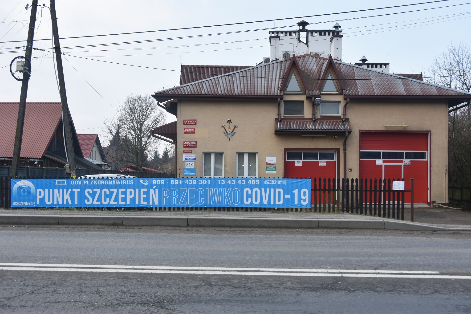 Budynek straży pożarnej z jasną elewacją, z dużymi drzwiami koloru czerwonego. Na ogrodzeniu przed budynkiem rozwieszony duży plakat koloru niebieskiego dot. szczepień.