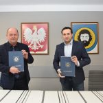 Dwóch mężczyzn stoi w jasnym pomieszczeniu. W rękach trzymają teczki granatowe. Za nimi na scianie wiszą obrazy herbu miasta i godła Polski.