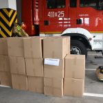 Kilkanaście pudełek stoi w hali strazy pożarnej. Za nimi wóz strazacki.