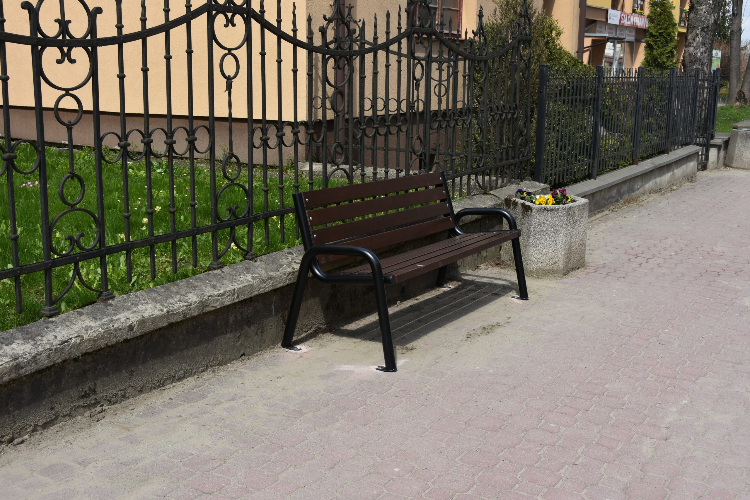 Ławka drewniana stoi na chodniku. Za nią ciemne metalowe ogrodzenie. Obok kwietnik.